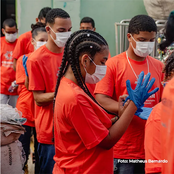Jovens de camiseta laranja usam luvas durante a atividade prática